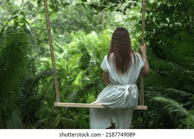 A Young Girl In A White Dress Rides On A Rope Swing In The Wood. The Concept Of Outdoor Recreation