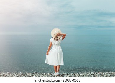 A young girl in a white dress stands on the seashore, looking into the distance. Holds the straw hat with his right hand