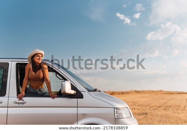 Young girl with white hat leaning out the window of\
her camper van leaning on the frame and watching the countryside\
landscape of a road trip from a frontal position. Concept of van\
life.