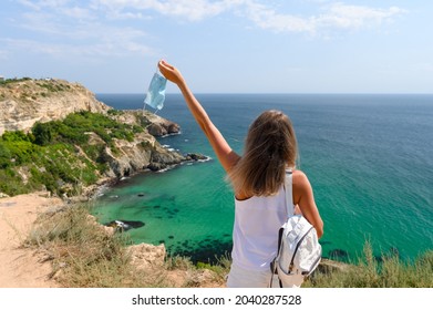 A young girl in a white T-shirt and a white backpack stands on the edge of a mountain with a medical mask in her hand and looks at the sea. - Powered by Shutterstock