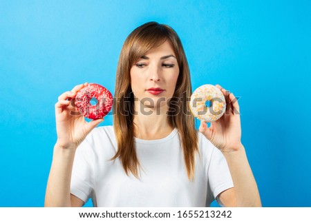young girl in a white T-shirt, holds two donuts and looks at them, on a blue background. Banner. Good mood, diet concept, bright colors