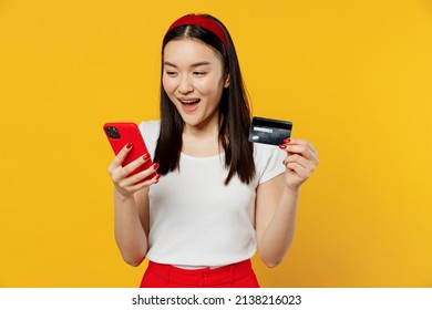 Young Girl Woman Of Asian Ethnicity 20s Years Old Wears White T-shirt Use Mobile Cell Phone Hold Credit Bank Card Do Online Shopping Order Delivery Isolated On Plain Yellow Background Studio Portrait