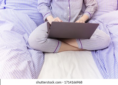 Young girl is working at laptop in bed, she is dressed in pajamas with blue stripes. Sunny morning in bedroom.
