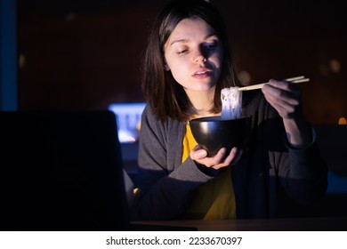 Young girl works at night at home with laptop and eats noodles, asian food with chopsticks. A woman watches a show, a movie and has dinner with ordered food from a cafe. - Shutterstock ID 2233670397