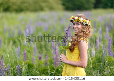 Young girl in yellow dress walking on meadow with lilac lupines. Warm summer day. Flower wreath for head