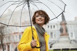 Young Girl In Yellow Raincoat With Transparent Umbrella In City. Young Beautiful Woman Wearing Rain Outfit And Eyeglasses While Walking On Street. Concept Of Modern Woman Lifestyle At Autumn.