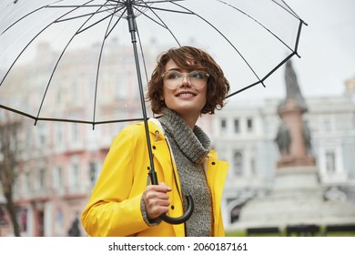 Young girl in yellow raincoat with transparent umbrella in city. Young beautiful woman wearing rain outfit and eyeglasses while walking on street. Concept of modern woman lifestyle at autumn. - Powered by Shutterstock