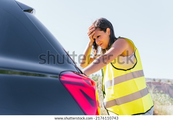 Young\
girl with yellow reflective vest leaning on the closed trunk of her\
broken down car and lamenting. Woman reflecting before placing the\
emergency warning systems for broken down\
vehicles