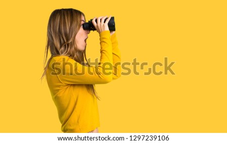 young girl with yellow sweater and looking in the distance with binoculars on isolated yellow background