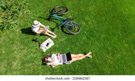Young girls with bicycle in park, using laptop computer and headphones, two student girls studying online outdoors sitting on grass near bike, aerial drone view from above