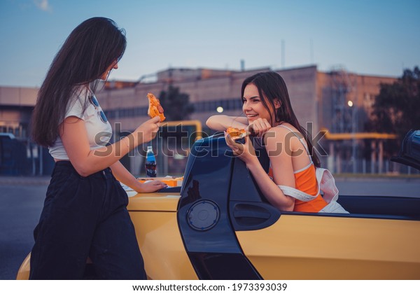 Young girls in casual outfit are smiling,\
eating pizza, posing in yellow car cabrio with french fries and\
soda in glass bottle on its trunk. Fast\
food