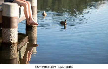 Young Girl's Legs Dangle From A Wooden Dock Above A Pond With Ducks Approaching In The Setting Sunlight