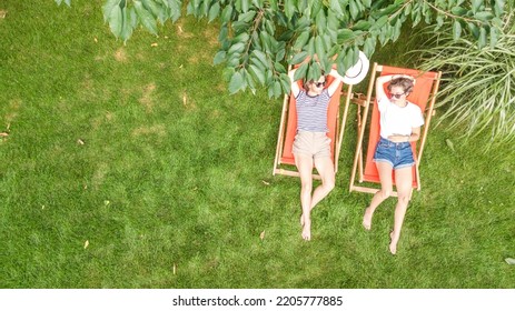 Young Girls Relax In Summer Garden In Sunbed Deckchairs On Grass, Women Friends Have Fun Outdoors In Green Park On Weekend, Aerial Top View From Above