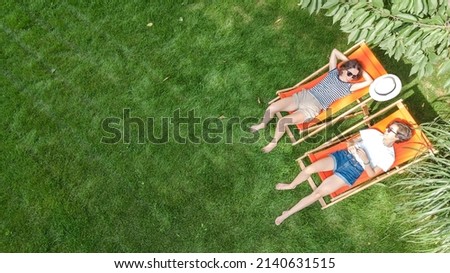 Young girls relax in summer garden in sunbed deckchairs on grass, women friends have fun outdoors in green park on weekend, aerial top view from above