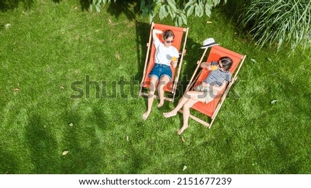 Young girls relax in summer garden in sunbed deckchairs on grass, women friends have fun outdoors in green park on weekend, aerial top view from above
