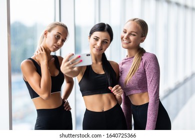 Young Girls In Sportswear Taking Selfie In Gym. Three Female Friends In Healthclub Taking Selfie With Mobile Phone.