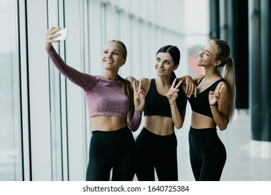 Young Girls In Sportswear Taking Selfie In Gym. Three Female Friends In Healthclub Taking Selfie With Mobile Phone.