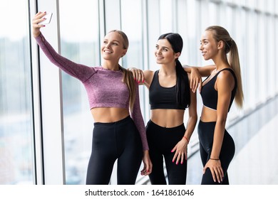 Young Girls In Sportswear Taking Selfie In Gym. Three Female Friends In Healthclub Taking Selfie With Mobile Phone.