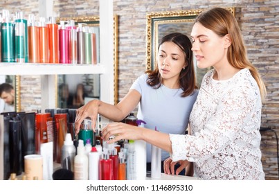 Young Glad Cheerful  Women Attentively Looking Hair Care Products At Beauty Salon