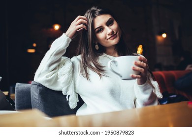 Young glad female in casual outfit looking at camera sitting in comfortable armchair touching hair and drinking hot coffee in cafe - Powered by Shutterstock