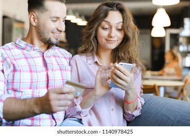 Young gladden wife and husband using smartphone and debit card at cafe. Concept of purchasing online and shopping, modern technology.