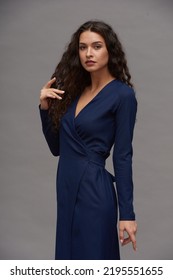 Young Glamorous Female Fashion Model In Navy Blue Smart Casual Dress Touching Her Dark Long Wavy Hair While Looking At Camera