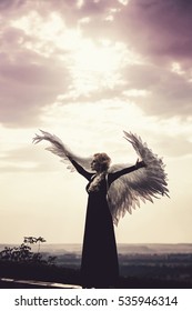 Young Glamorous Woman In A Black Dress With A Large White Angel Wings On The Background Of A Dramatic Sunset Sky. The Concept Of A Fallen Dark Angel