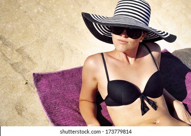 Young Glamorous Woman In A Summer Hat, Sunglasses And A Black Bikini Laying On The Beach.