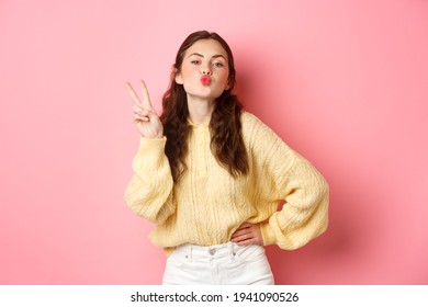 Young Glamour Woman Pucker Her Lips, Making Kissing Face And V-sign, Show Peace Gesture, Standing Against Pink Background