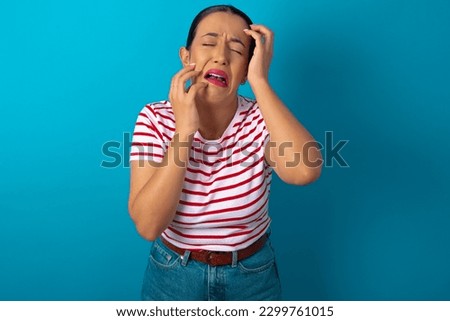 Young gloomy beautiful woman wearing striped T-shirt over blue studio background, hiding face with hands pouting and crying, standing upset and depressed complaining about job problem.