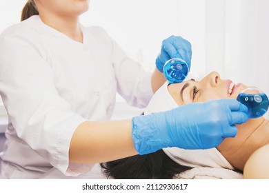 Young Good Looking Brunette Woman In Beauty Salon During Treatment Massage With Blue Cold Glass Ice Balls. Cryo Therapy. Face And Skin Care.