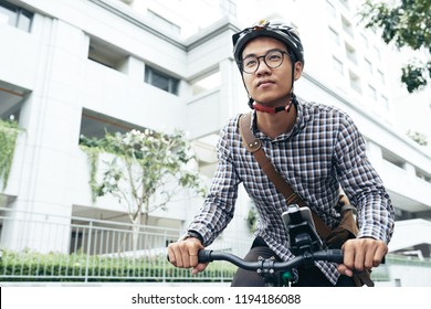 Young Good-looking Asian Man Wearing Safety Helmet And Glasses Looking Forward While Riding Bicycle To Work