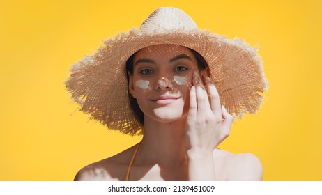 Young good-looking brunette Caucasian woman in a straw hat puts spf cream on her cheeks, enjoys the sun and smiles for the camera against yellow background | Spf cream concept