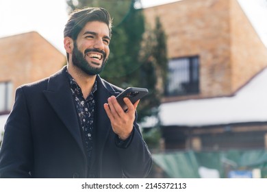 A Young Good-looking Latin American Man In Black Clothes Holding A Phone And Smiling Widely Medium Closeup Outdoor Urban Background . High Quality Photo