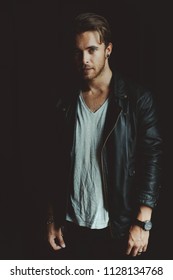 Young good-looking man with perfect hairstyle, and a stud earring, wearing black leather jacket posing in the darkness of the room, lit by daylight. Looking at camera. Texture effect added.