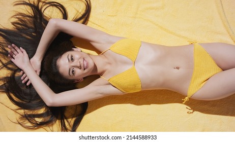 Young good-looking slim brunette Asian woman with long flowing hair in yellow knitted bikini lies on yellow terry blanket sunbathing, putting hands up on hair and smiling wide | Sun screen commercial