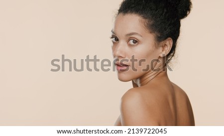 Young gorgeous African American woman with bun hairstyle turns and looks over her shoulder on beige background | Beauty care commercial concept