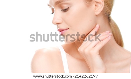 Young gorgeous Caucasian woman with golden hair in white crop top touches her jaw line turning her head aside on white background | Perfect skin for beauty commercial