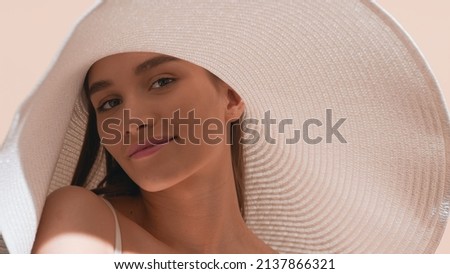 Young gorgeous dark-haired European woman in a big white hat with a smear of sunscreen on her cheek lifts up her shoulder and looks at camera against beige background | Sunscreen concept