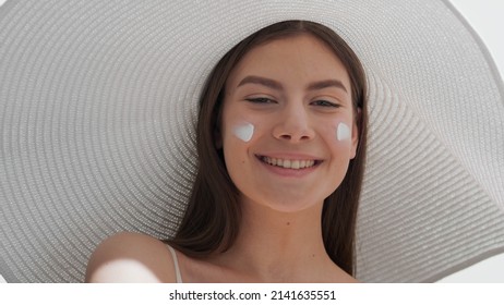 Young Gorgeous European Woman With Brown Hair In A Big White Hat And Two Smears Of Sun Cream On Her Cheeks Looks At Camera And Smiles Widely Against White Background | Sunscreen Application