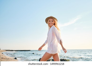 Young gorgeous smiling blond woman in white shirt and hat happily walking by the sea