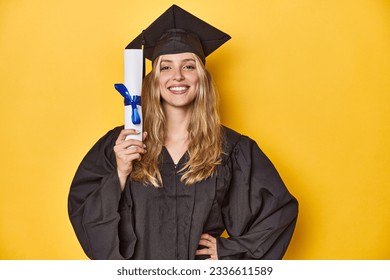 Young graduate woman in cap and gown, holding a diploma on a yellow studio background.