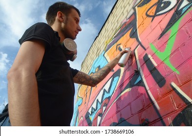 Young graffiti artist with backpack and gas mask on his neck paints colorful graffiti in pink tones on brick wall. Street art and contemporary painting process