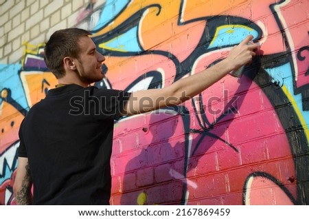 Young graffiti artist with backpack and gas mask on his neck paints colorful graffiti in pink tones on brick wall. Street art and contemporary painting process. Entertainment in youth subculture