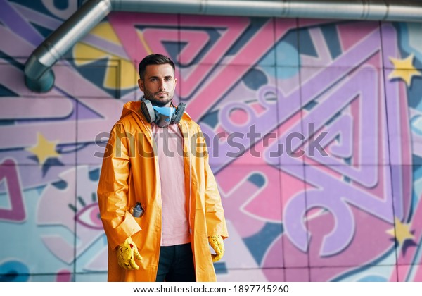 Young graffiti painter\
standing near the wall with his paintings with copy space. Street\
art concept