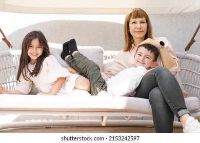 Young Grandmother With Smiling Grandchildren Sitting On Swing Bench In Pergola On A Sunny Day.