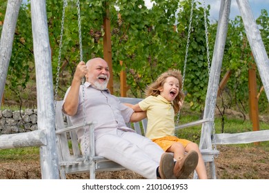 Young Grandson And Old Grandfather Swinging In Spring Garden. Grand Dad And Grandson Sitting On Swing In Park.