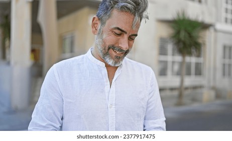 Young grey-haired hispanic man, confidently smiling, standing, basking in sunny street ambiance, a cool look in his eyes, exuding positivity.