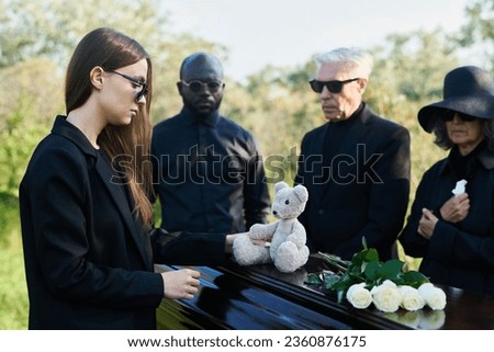 Young grieving woman in mourning attire and sunglasses putting grey teddybear on top of closed lid of coffin with dead body of her child