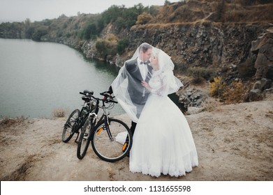 A Young Groom In A Black Suit And A Beautiful Bride In A Lace Dress With A Long Veil Stand Near A Bicycle Under A Veil And Laugh At The Background Of Rocks And Stones. Autumn Wedding Portrait Of Gay.